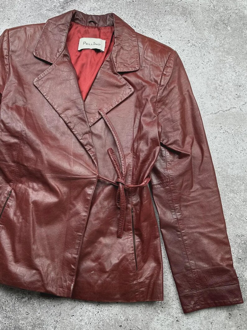 May include: A burgundy leather jacket with a tie belt closure. The jacket has a notched collar and a red satin lining. The brand name "Pellino" is visible on a tag inside the jacket.