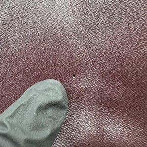 K&ouml;nnte beinhalten: Nahaufnahme einer strukturierten, weinroten Lederoberfl&auml;che mit einem kleinen Loch. Ein dunkelgrauer Handschuh ist teilweise in der linken unteren Ecke zu sehen, der das Leder ber&uuml;hrt. Das Leder hat ein subtiles, k&ouml;rniges Muster.