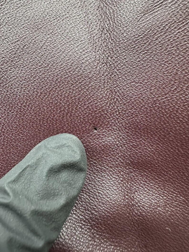 K&ouml;nnte beinhalten: Nahaufnahme einer strukturierten, weinroten Lederoberfl&auml;che mit einem kleinen Loch. Ein dunkelgrauer Handschuh ist teilweise in der linken unteren Ecke zu sehen, der das Leder ber&uuml;hrt. Das Leder hat ein subtiles, k&ouml;rniges Muster.