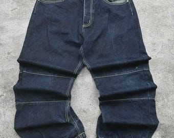 May include: Dark wash denim jeans with a unique design. Features include contrasting stitching, a patch on the front pocket, and a dark patch detail on the front. The jeans have a relaxed fit and a distinctive seam design on the legs.