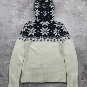 May include: A hooded sweater in cream and navy blue. The hood and upper chest feature a navy blue pattern of white snowflakes. The lower portion of the sweater is cream-colored. The sleeves have ribbed cuffs. The sweater is made of a soft, knitted material.