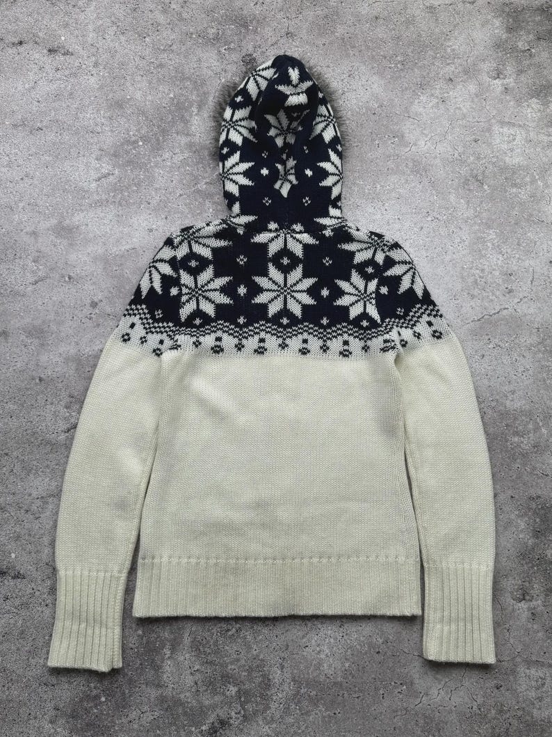 May include: A hooded sweater in cream and navy blue. The hood and upper chest feature a navy blue pattern of white snowflakes. The lower portion of the sweater is cream-colored. The sleeves have ribbed cuffs. The sweater is made of a soft, knitted material.