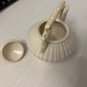 Op de afbeelding: Crèmekleurige keramische theepot met een geribbelde body en een textuur handvat. Het deksel ligt ernaast. De theepot heeft een tuit en een zeefje binnenin. De theepot staat op een wit oppervlak.
