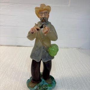 Pode incluir: Estatueta de um homem idoso usando chapéu, casaco e carregando uma bolsa verde. O homem está segurando um pequeno objeto nas mãos. A estatueta está em uma base azul e verde.