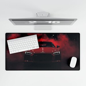 May include: A black rectangular desk mat featuring a red sports car design with red smoke. A white keyboard and mouse are on the mat. A computer monitor is above the mat.