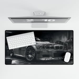 May include: A black and white photo of a silver car in a garage. The car is a BMW M3 with a black interior and a black steering wheel. The car is sitting on a black mat with a white keyboard and a white mouse on the mat.