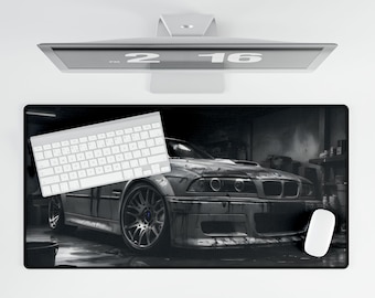 May include: A black and white photo of a silver car in a garage. The car is a BMW M3 with a black interior and a black steering wheel. The car is sitting on a black mat with a white keyboard and a white mouse on the mat.