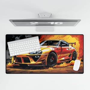 May include: A rectangular desk mat featuring a stylized illustration of a yellow and red sports car with black wheels, set against a fiery background. A white keyboard and mouse are on the mat. A computer monitor is above the mat.