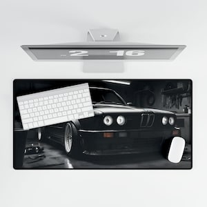 May include: A black and white photo of a black BMW car in a garage. The car is facing the camera and the headlights are on. The car is sitting on a black surface. A white keyboard and a white mouse are also in the photo.