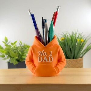 以下が含まれることがあります： オレンジ色のパーカー型ペン立て。前面に「No. 1 DAD」の刺繍入り。様々な色のペンや鉛筆が入っています。ユニークなデスクアクセサリーです。