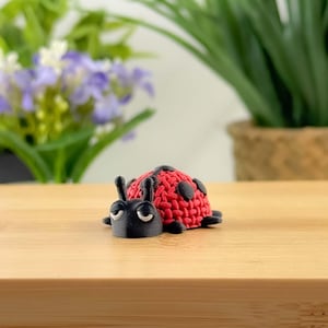 May include: A small ladybug ornament. The ladybug has a red, textured body with black spots and a black head with curved white details. The ornament is on a light brown surface.