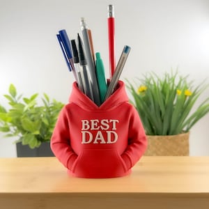 以下が含まれることがあります： 「BEST DAD」の白い文字が入った赤いパーカー型のデスクオーガナイザー。赤い鉛筆、緑のペン、青と黒のペンなど、さまざまなペンや鉛筆が入っています。木製の表面に置かれています。