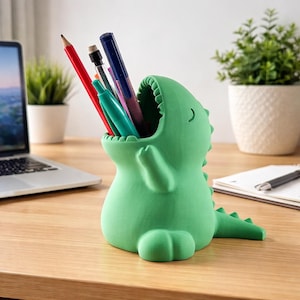 Könnte beinhalten: Ein grüner, dinosaurierförmiger Schreibtisch-Organizer mit Stiften und Bleistiften. Der Organizer hat ein offenes Munddesign und einen Schwanz. Der Schreibtisch-Organizer besteht aus Kunststoff und steht auf einem Holzschreibtisch.