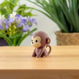 Può includere: Piccola statuetta di scimmia marrone con viso, orecchie e pancia color beige chiaro. La scimmia è seduta su una superficie di legno. Lo sfondo comprende piante verdi e fiori viola.