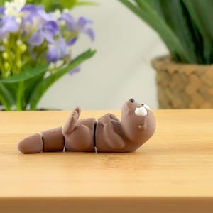 May include: A brown, segmented otter figurine lying on its back with its arms raised. The otter has white eyes and is positioned on a wooden surface. The figurine is a whimsical decorative item.