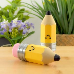 Pode incluir: Dois objetos amarelos em forma de lápis com pontas pretas e rostos sorridentes. Um lápis está em pé e o outro está deitado. Ambos têm uma borracha rosa e faixas de metal prateado. O fundo inclui plantas verdes e uma superfície de madeira.