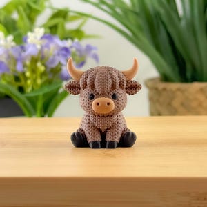 Op de afbeelding: Een bruin Highland-koebeeldje met een gebreide textuur. Het beeldje heeft bruine hoorns en een lichtbruine snuit. De koe zit met zijn poten onder zijn lichaam. De achtergrond bevat groene planten en paarse bloemen.