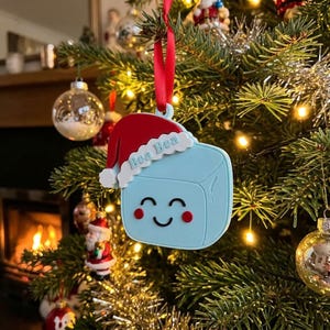 May include: A light blue square-shaped ornament with a smiling face, red cheeks, and a red Santa hat with white trim. The name Bea Bea is embossed in light blue on the white trim of the hat. The ornament hangs from a red ribbon.