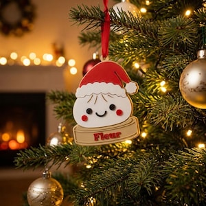 May include: A white dumpling-shaped Christmas ornament wearing a red Santa hat with a white trim. The ornament features a smiling face with black eyes and red cheeks, resting on a tan base with the name Fleur written in red capital letters.