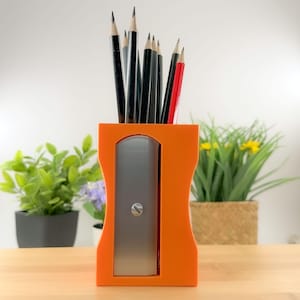 May include: An orange pencil sharpener with a silver blade and a collection of pencils. The pencils are black and red, with sharpened tips. The sharpener is on a wooden surface.