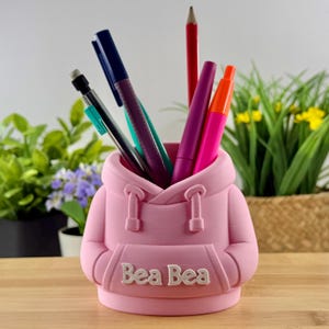 May include: A pink pen holder in the shape of a hoodie, with the text "Bea Bea" on the front. It holds pens and pencils in blue, purple, red, and orange. The pen holder is on a wooden surface, with greenery in the background.