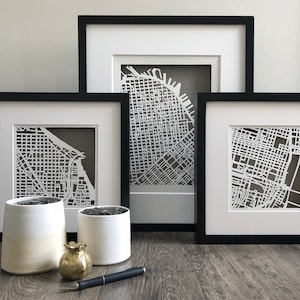 May include: Three black framed prints of city maps with white paper cutouts. The prints are stacked on a wooden surface with two white ceramic pots and a gold decorative object.