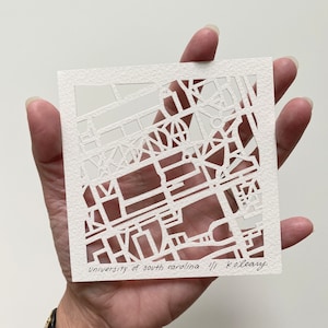 May include: White paper cut-out art of a map of the University of South Carolina campus. The artist's signature is in the bottom right corner: "University of South Carolina // K. O'Leary."