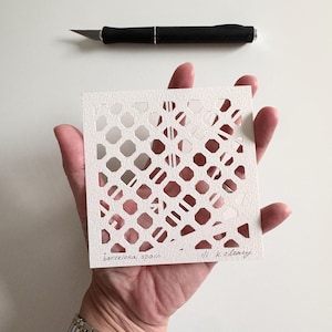 May include: A square white paper cutout featuring a geometric lattice pattern of diamond and octagonal shapes. The bottom left corner is inscribed with the text 'barcelona, spain' and the bottom right with '1/1 k. o'leary'. A black craft knife is positioned above.