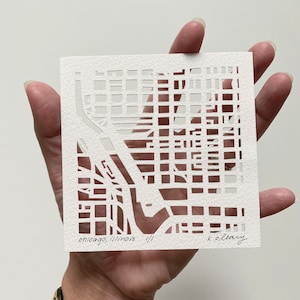 May include: A square, white paper cut-out depicting a map of Chicago, Illinois. The intricate design shows streets and buildings. The text "chicago, illinois 1/1 k.ollary" is handwritten at the bottom.