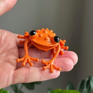 May include: An orange frog figurine with black eyes and a textured back. The frog has four legs and is posed in a sitting position. The figurine is made of a glossy material and is a vibrant orange color.