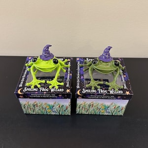 May include: Two Smiling Frog Wizard figurines, one lime green and one olive green, each wearing a small purple wizard hat with gold stars. The frogs are perched on top of their respective square boxes, which feature a floral and starry design.