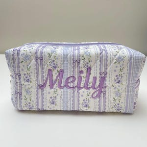 May include: A rectangular quilted cosmetic bag featuring a white and light purple floral pattern with vertical stripes and bows. The name Meily is embroidered in purple script across the center front. The bag has a top zipper closure.
