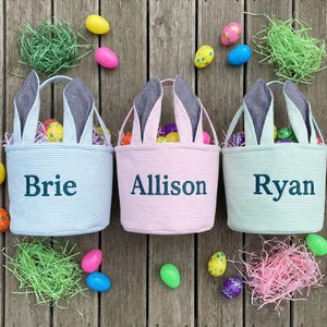 May include: Three Easter baskets in blue, pink, and green, each with bunny ears and a name: Brie, Allison, and Ryan. The baskets are filled with colorful eggs. Easter grass and eggs in various colors surround the baskets.
