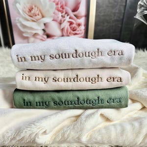 May include: Three sweatshirts stacked on top of each other. The top sweatshirt is gray, the middle sweatshirt is beige, and the bottom sweatshirt is green. All three sweatshirts have the same embroidered text: "in my sourdough era".