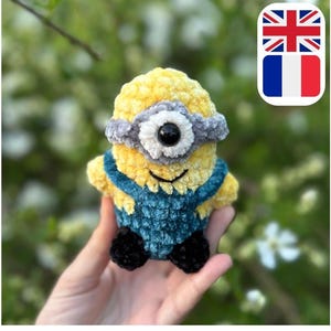 Könnte beinhalten: Ein handgefertigtes, gehäkeltes Minion-Spielzeug in Gelb und Blau. Der Minion hat ein Auge, ein graues Stirnband und blaue Latzhosen. Das Spielzeug wird in einer Hand vor einem verschwommenen grün-weißen Hintergrund gehalten.