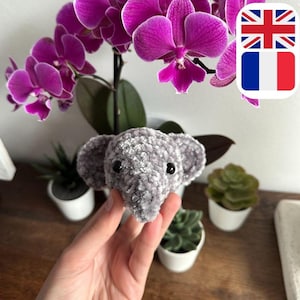 Peut inclure: Un éléphant en peluche gris crocheté avec des yeux noirs. Le jouet est tenu dans une main avec une plante d'orchidée rose en arrière-plan.