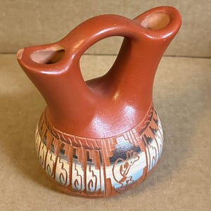 May include: A terracotta-colored clay vessel featuring a double-spout design connected by a central handle. The rounded base is decorated with etched geometric patterns and a stylized figure in white, black, and blue tones against the clay surface.