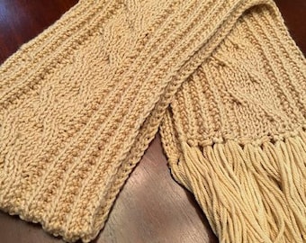 Puede incluir: Una bufanda de punto beige con un patrón de punto de cable y flecos en los extremos.