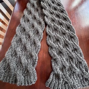 May include: A grey and white knitted scarf with a cable knit pattern. The scarf is made from a soft, textured yarn and has a thick, warm appearance. The scarf is displayed on a wooden surface.