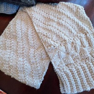 May include: A cream-coloured knitted scarf with a textured pattern. The scarf is folded in half and the ends are overlapping.