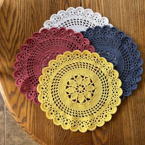 May include: A set of four circular crochet doilies in white, dark red, navy blue, and yellow. Each doily features a central floral pattern surrounded by intricate openwork stitching and a scalloped edge, layered to show the distinct colors.