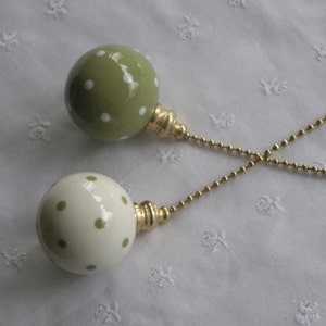 May include: Two spherical ceramic light pulls with gold-toned metal connectors and beaded chains. One pull is sage green with white polka dots, and the other is cream with sage green polka dots. The smooth, glossy finish reflects light.