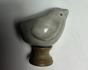 May include: A smooth, glossy, gray ceramic bird figurine perched atop a small, cylindrical, brown wooden base. The bird has a rounded body, a small pointed beak, and a dark, simple eye detail, creating a compact and solid decorative object.