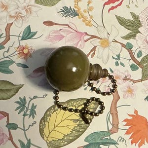 May include: A smooth, olive green spherical pull chain knob with a glossy finish, featuring a tiered, stepped neck design and a metallic bronze-toned ball chain connector attached to the base for use with ceiling fans or light fixtures.
