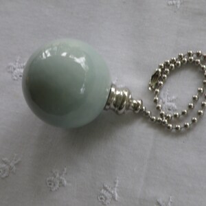 May include: A light green, spherical ceiling fan pull chain with a silver-colored metal base and a beaded chain. The pull chain is resting on a white, textured surface.