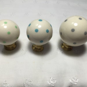 May include: Three round, white ceramic cabinet knobs with brass bases. Each knob is decorated with a pattern of colorful polka dots in green, blue, and gray. The knobs are displayed on a white, textured surface.