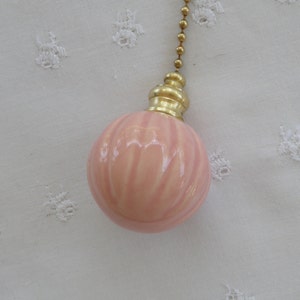 May include: A light pink, ribbed ceramic light pull with a gold-colored metal cap and a brass ball chain. The pull has a spherical shape and is set against a white, textured background.