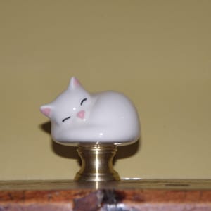 May include: A white ceramic cat figurine with pink accents, sleeping peacefully. The cat is mounted on a brass base. The figurine is a decorative item, possibly a cabinet knob or drawer pull.