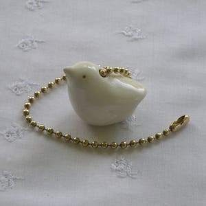 May include: A cream-colored ceramic bird-shaped pull chain for a ceiling fan or light fixture. The bird is attached to a gold-colored ball chain with a matching clasp. The bird is sitting on a white, textured surface.