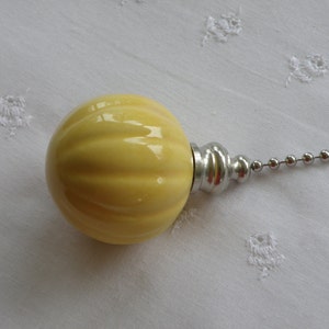 May include: A light yellow, ribbed ceramic light pull with a silver-toned metal base and a beaded chain. The pull is spherical and has a glossy finish. The chain is made of small, round metal beads.